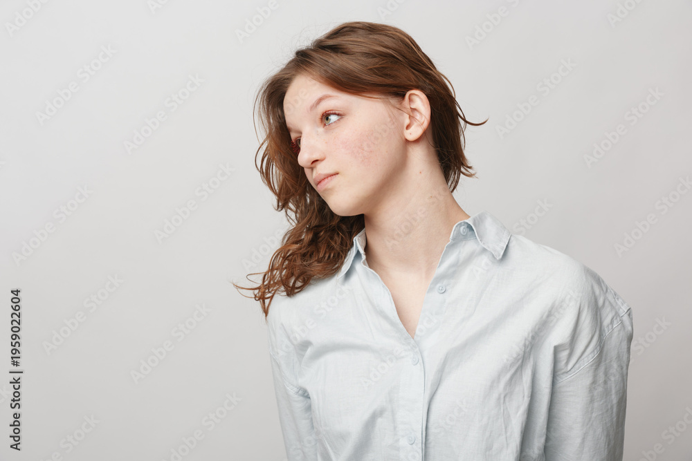 Obraz premium Young woman with curly brown hair wearing a white shirt looks sideways against a plain light background. Concept of natural beauty, casual style, and thoughtful mood in studio.