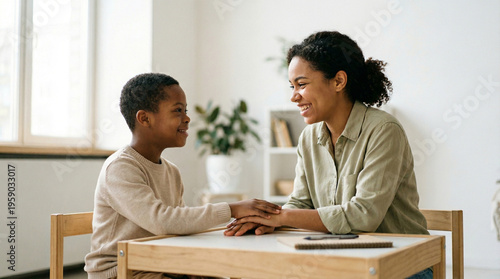 Black mother and young son with Down syndrome hold hands and talk warmly at a table, sharing a nurturing educational moment at home, family love, inclusive growth.