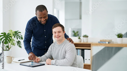 A black male mentor stands by a young man with Down syndrome who is using a laptop in a bright office. inclusive workplace, professional growth, mentorship, corporate diversity.