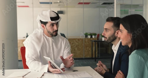 Happy young Caucasian couple consulting male Arabic real estate seller, watching virtual tour, property offer on tablet, discussing buying or rent legal terms, mortgage, investment