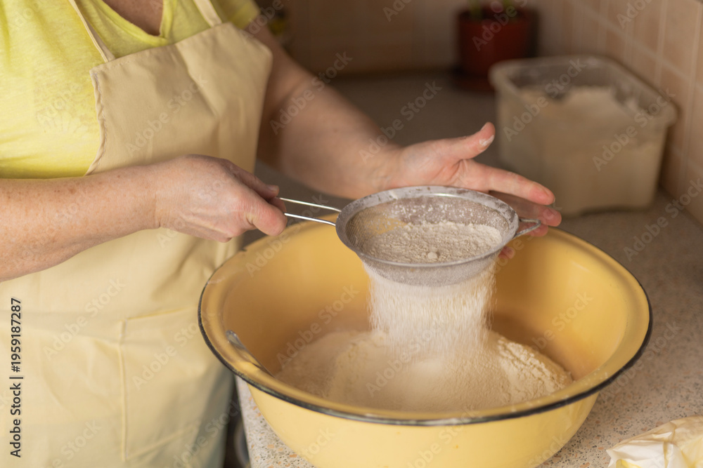 Fototapeta premium Adult woman hands sifting flour over bowl with sieve in kitchen