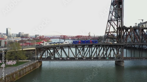 4K aerial drone view of MAX light rail train crossing Steel Bridge with station in downtown Portland Oregon USA over Willamette River in sunrise light. Urban transit and transportation scene.