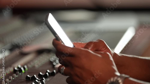 Close-up of hands holding smartphone with audio mixing console in background. Recording studio, music production, sound engineer using mobile phone at soundboard. Modern technology in music studio.