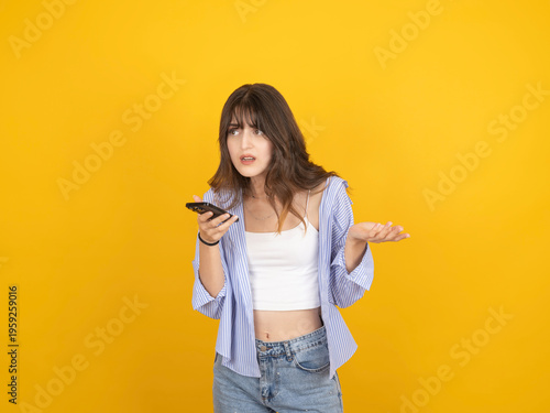 Confused young woman holding smartphone with questioning gesture, wearing striped shirt and denim, standing against bright yellow studio background with copy space. Problem and uncertainty concept.