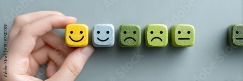 A person picks a cheerful yellow smiley face block from a row of wooden emotion cubes on a smooth grey surface symbolizing positive feedback customer satisfaction or choice