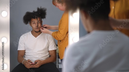Hairdresser masterfully braids an african american man's afro hair. The man sits in a chair, watching his reflection in a mirror