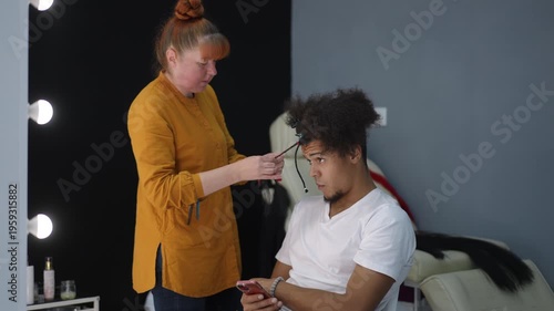 Young black man sitting in a barber chair having his hair braided by a professional stylist while checking his smartphone in a salon