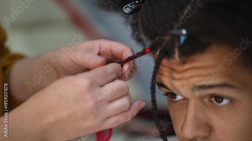 Close-up of a stylist's hands braiding colorful synthetic hair into the dreadlocks of a male client. Afro-american man getting a new hairstyle at a beauty salon