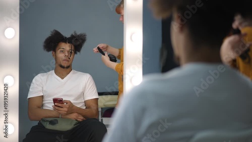 Young biracial man sits in a makeup chair while a hairdresser styles his afro hair. The actor is using his smartphone backstage