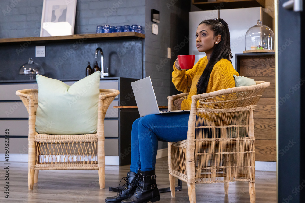 Fototapeta premium Adult Asian female sitting in kitchen wearing yellow sweater holding laptop and red mug, copy space