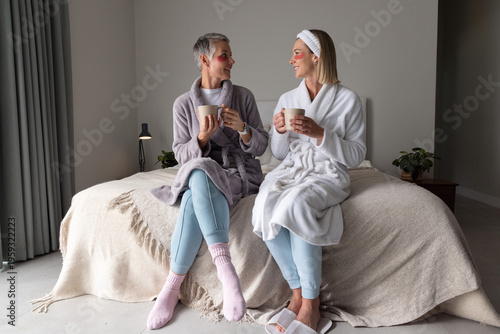 Senior mother and daughter sitting on bed in bedroom, wearing robes and eye patches, sipping mugs