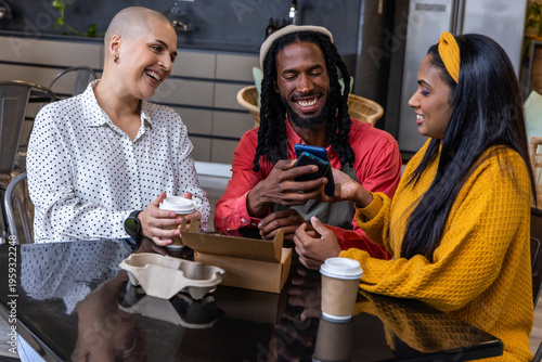 Diverse friends sitting at cafe table, laughing, sharing smartphone, coffee cups, takeout