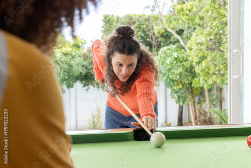 Female friends playing on pool table at home with woman in orange sweater aiming wooden cue