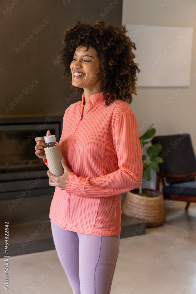 Fototapeta premium African American woman smiling, holding textured bottle at home wearing coral jacket near fireplace