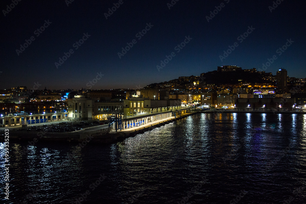 Obraz premium Panorama of Naples, Italy, at night. The city lights of a coastal port glitter against the dark night sky, reflecting beautifully on the calm sea surface. There are buildings and a distant hill.
