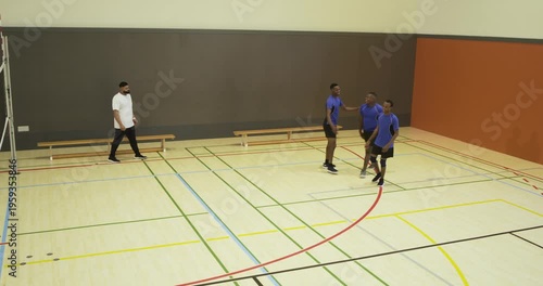 Blue-shirted males moving in gym as ball rolling, huddling with coach checking spots, copy space