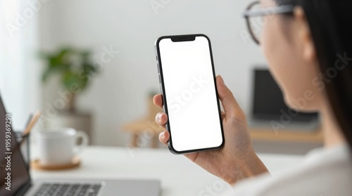 Person holds a smartphone while sitting at a desk with a laptop and plant in the background