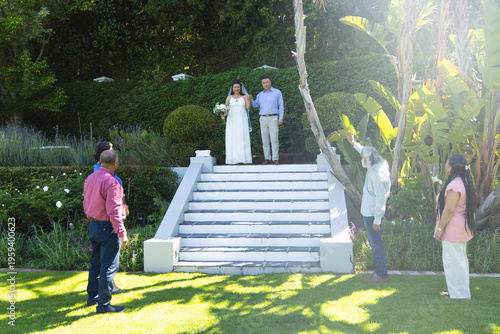 Posing bride and groom on white steps at garden, guest raising smartphone capturing wedding bouquet