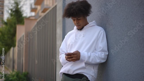 Young african american man with an afro hairstyle leaning against a wall and using a smartphone. He is wearing wireless earphones