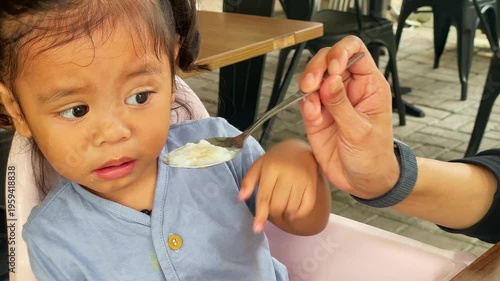 Little girl grimaces and refuses to eat the porridge that her mom feeds her.
