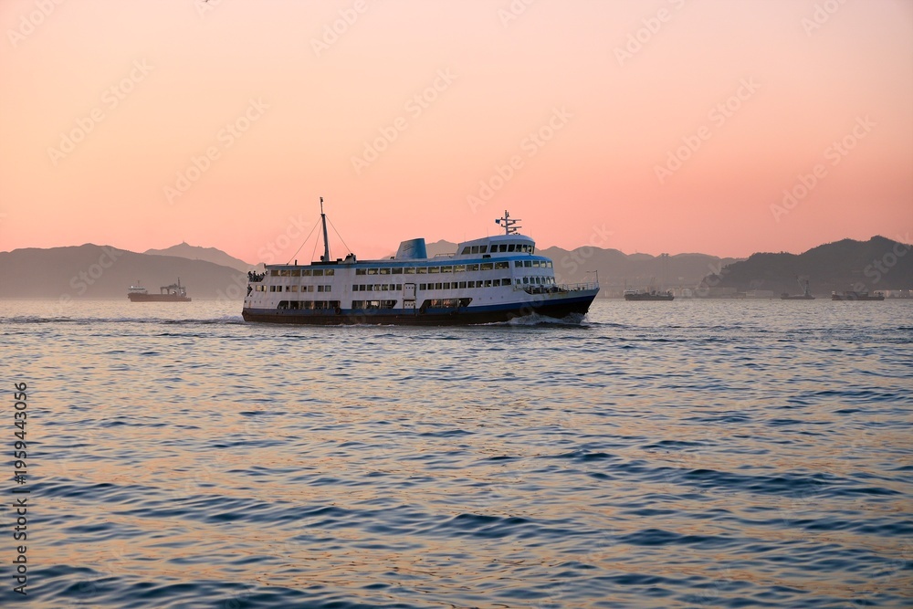 Obraz premium A classic white and blue Star Ferry sails across the water as the sky turns pink behind the distant mountain silhouettes