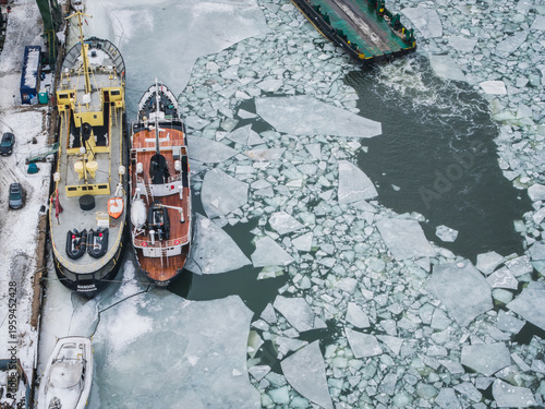 Aerial view of ships docked beside a cold, ice-covered canal, the stark white and grey ice contrasting with the ships' warm hues, Gdansk, Poland.