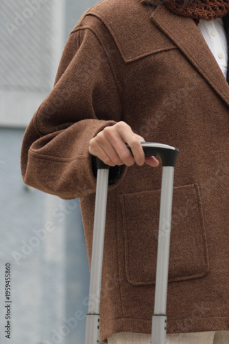 Close-up of a woman in a brown wool coat holding the handle of a suitcase. The shot expresses calm readiness, autumn travel mood, and refined minimalism in neutral tones.