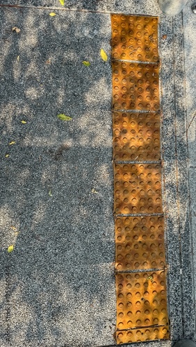 Downward perspective of weathered yellow tactile paving tiles set into a grey aggregate sidewalk under dappled sunlight.