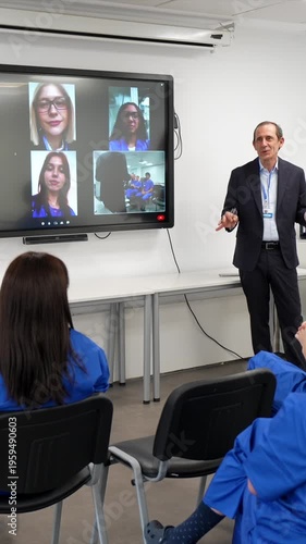 Male doctor leading a medical seminar. Teaching a class of students in scrubs while connecting with other participants remotely through a large video conference screen in a modern hospital classroom
