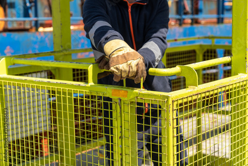 An industrial worker in protective gloves closes the safety latch on a metal work platform at a construction or port site. Workplace safety, equipment use, and industrial processes.