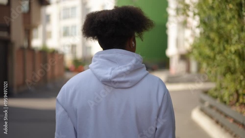 Rear view shot of a young black man with a unique hairstyle walking along an urban street. He is wearing a white hoodie and wireless earbuds