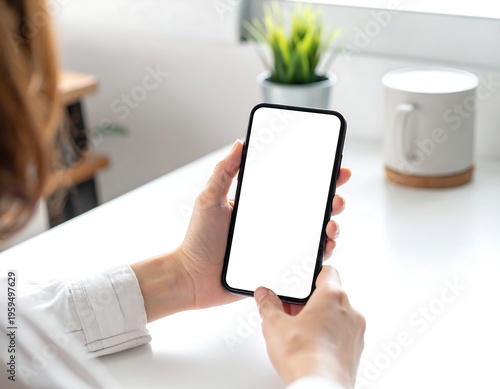 A person holds a blank-screened handheld device with two hands, resting atop a white table beside a decorative plant and mug