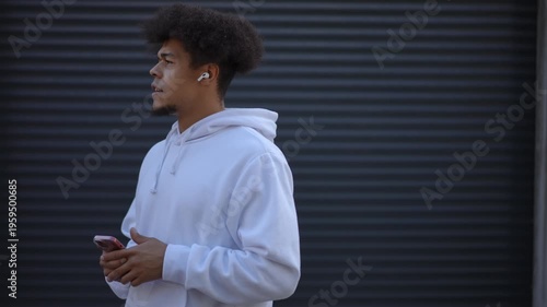 Side view of a young mixed-race man with an afro hairstyle having a pleasant conversation using his smartphone and wireless earphones outdoors