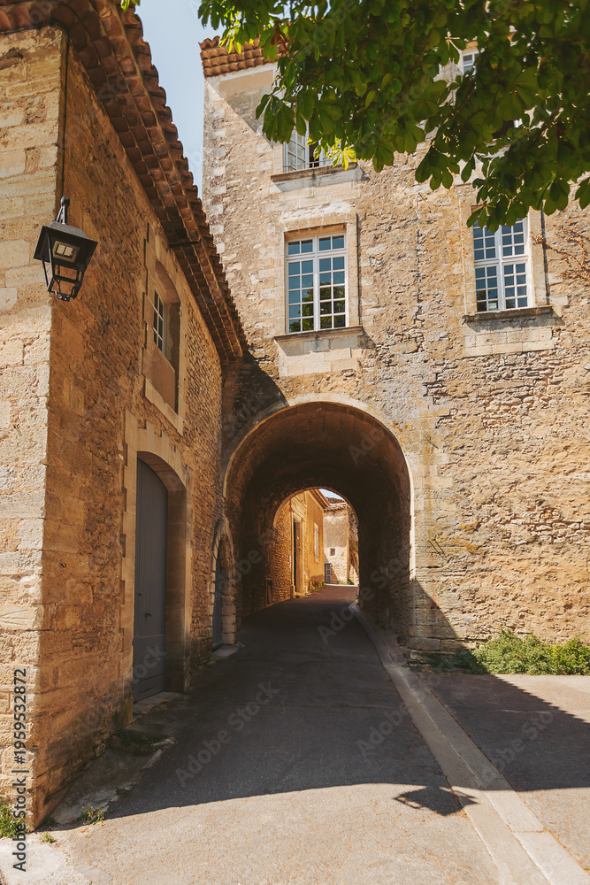 Fototapeta premium A picturesque stone archway frames a serene view of a narrow cobblestone street, illuminated by bright sunshine in a quaint French village. Old buildings whisper stories of the past