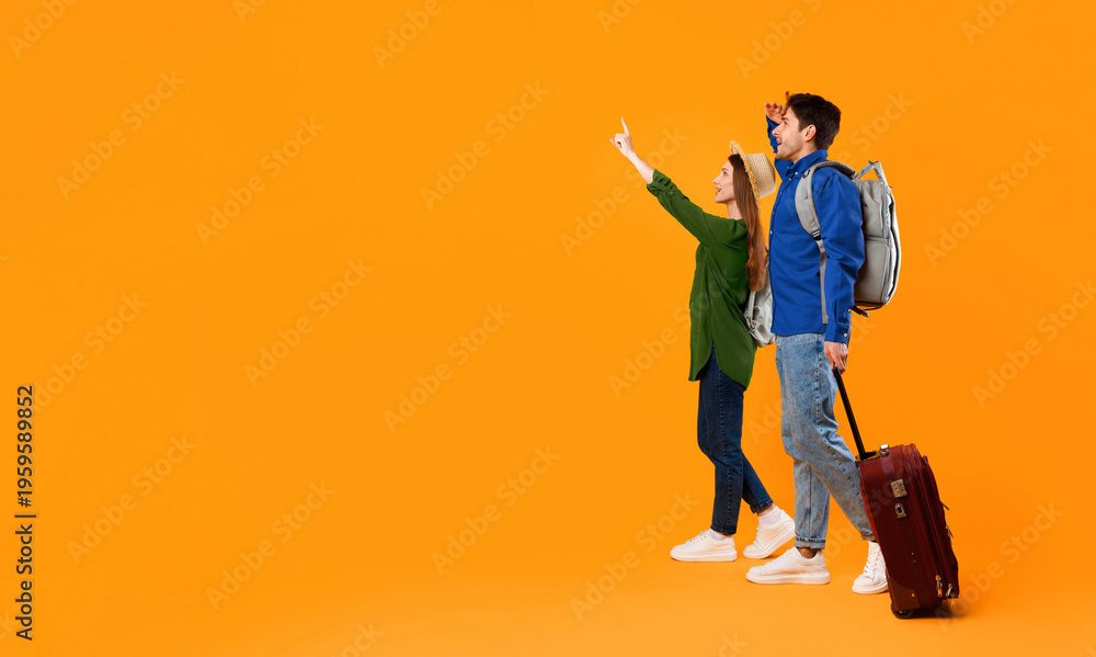 Fototapeta premium A couple stands on an orange background. They seem excited and point at something ahead. They carry a suitcase and have backpacks. Their expressions show anticipation for their journey.
