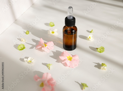 Organic essential oil bottle with glass dropper surrounded by spring flowers. Amber glass bottle with natural serum for skincare or aromatherapy on white surface with sunlight shadows.
