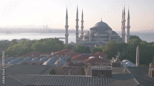 The Blue Mosque with tall minarets rises above rooftops and green trees with the Istanbul skyline and Bosphorus in the background. Soft sunrise light and warm tones create a peaceful and iconic