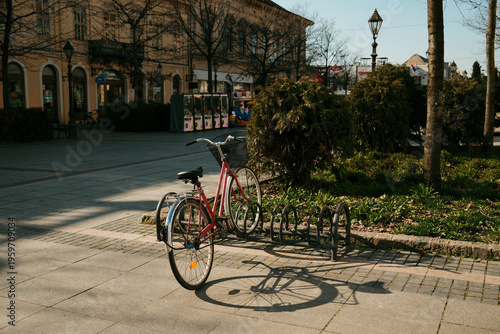 Red bicycle parked near rack in urban square with soft sunlight and shadows in Sombor, Serbia. Concept of sustainable transport, city lifestyle and eco friendly mobility
