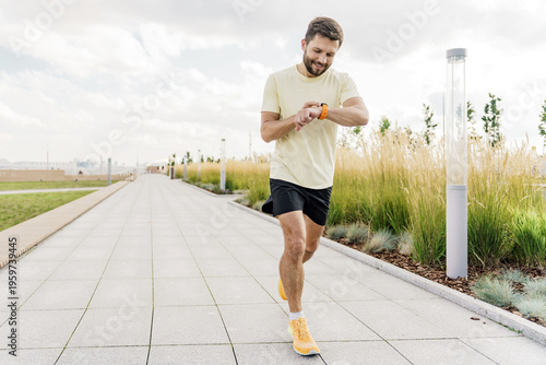 Bearded man running on a wide paved park path, checking his fitness data on a bright smartwatch under a cloudy sky