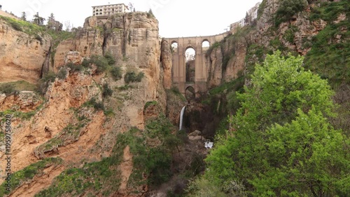Panoramic view of Puente Nuevo bridge in Ronda, featuring a waterfall at the bottom of El Tajo gorge surrounded by spring vegetation and cliffs.