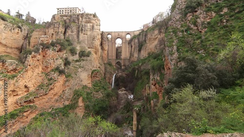 Wide shot of the historic Puente Nuevo bridge in Ronda, Andalusia, showing the dramatic Tajo gorge, a flowing waterfall, and steep limestone cliffs.