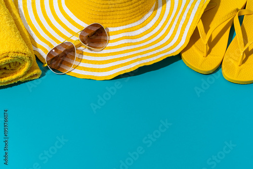 Bright yellow beach hat with stripes, paired with stylish sunglasses. Yellow beach accessories on turquoise blue background with copy space. Soft, rolled towel and vibrant yellow flip-flops.
