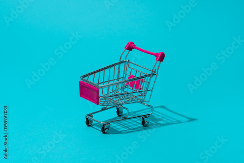 Minimalist photo of deep pink push cart as concept of supermarket, retail and sale season. Shopping trolley on turquoise blue background central composition.