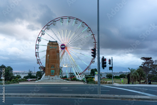 The iconic Sky Wheel at St Kilda Beach amusement park under a cloudy sky, featuring the historic Luna Park entrance and Melbourne city skyline in the background.