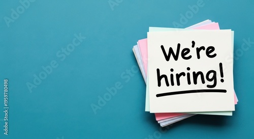 We're hiring message on sticky notes, recruitment announcement on paper stack, job vacancy concept on blue background with copy space