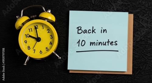 Yellow vintage alarm clock with back in 10 minutes note, blue sticky note on black background, time management and break concept