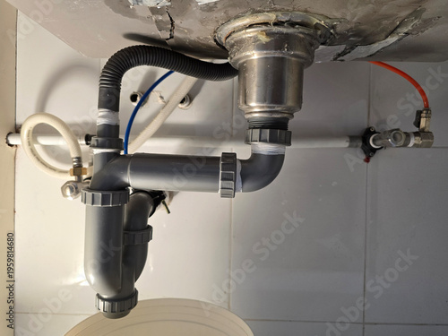 A view of the grey plastic P-trap drainage system and water supply hoses installed under a stainless steel kitchen sink.