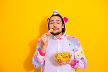 Happy man in colorful unicorn pajama eating chips in front of vibrant yellow background for fun lifestyle stock photo