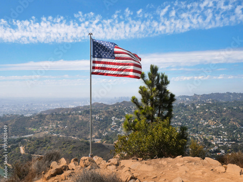 Flag of the United States of America waving on Cahuenga Peak at Wisdom Tree with the Hollywood Reservoir and skyline of LA in the background during a summer season in Los Angeles, California, USA