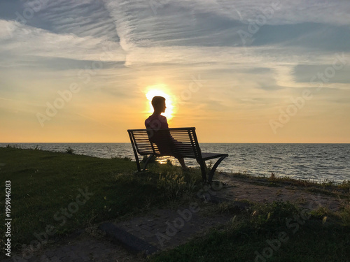 Silhouette of a man sitting on a bench, quietly watching a golden sunset over the sea. Warm light reflects across the water, creating a calm and timeless travel atmosphere, with a sense of solitude.
